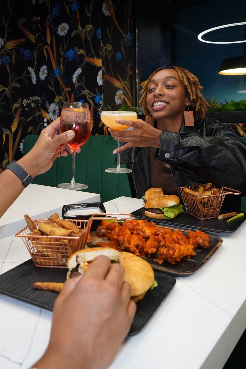 Woman with locs toasting cocktails at restaurant table with burgers, buffalo wings, and fries, showing social dining atmosphere with decorative wallpaper and modern lighting