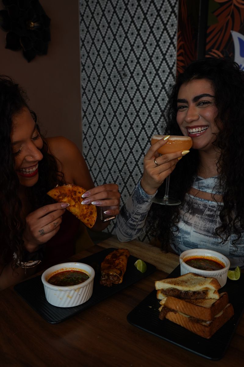 Two women at restaurant table - one holding pizza slice and another toasting with cocktail, with grilled sandwiches and soup bowls on dark plates, geometric patterned wall background showing fun social dining atmosphere