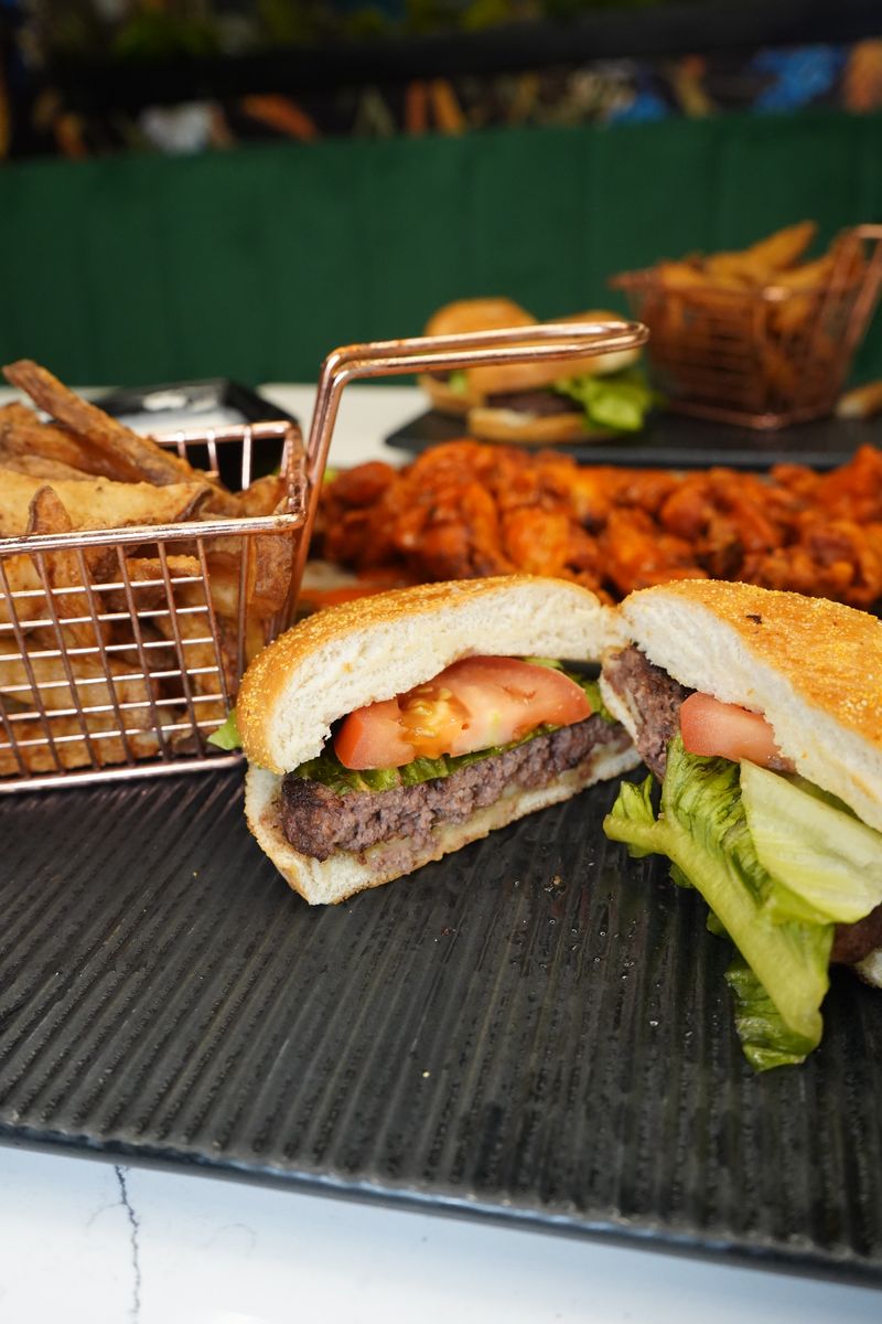 Professional food photography showing gourmet burger cut in half revealing layers of beef patty, tomato, lettuce on artisanal bun, served with seasoned fries in copper wire basket and buffalo wings in background, all on dark slate serving board