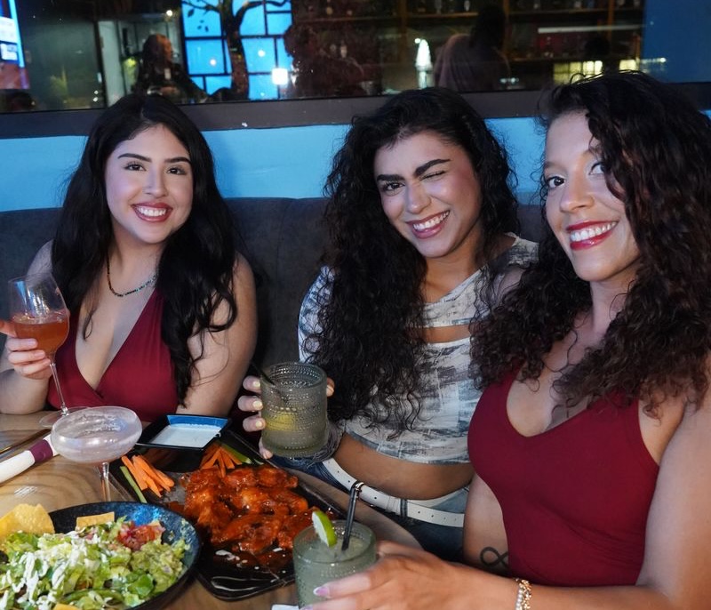 Three women enjoying social dining experience at restaurant booth with Carolina blue ambient lighting in background, wooden table with buffalo wings, salad, and cocktails, capturing the energetic Franklin Street game day atmosphere