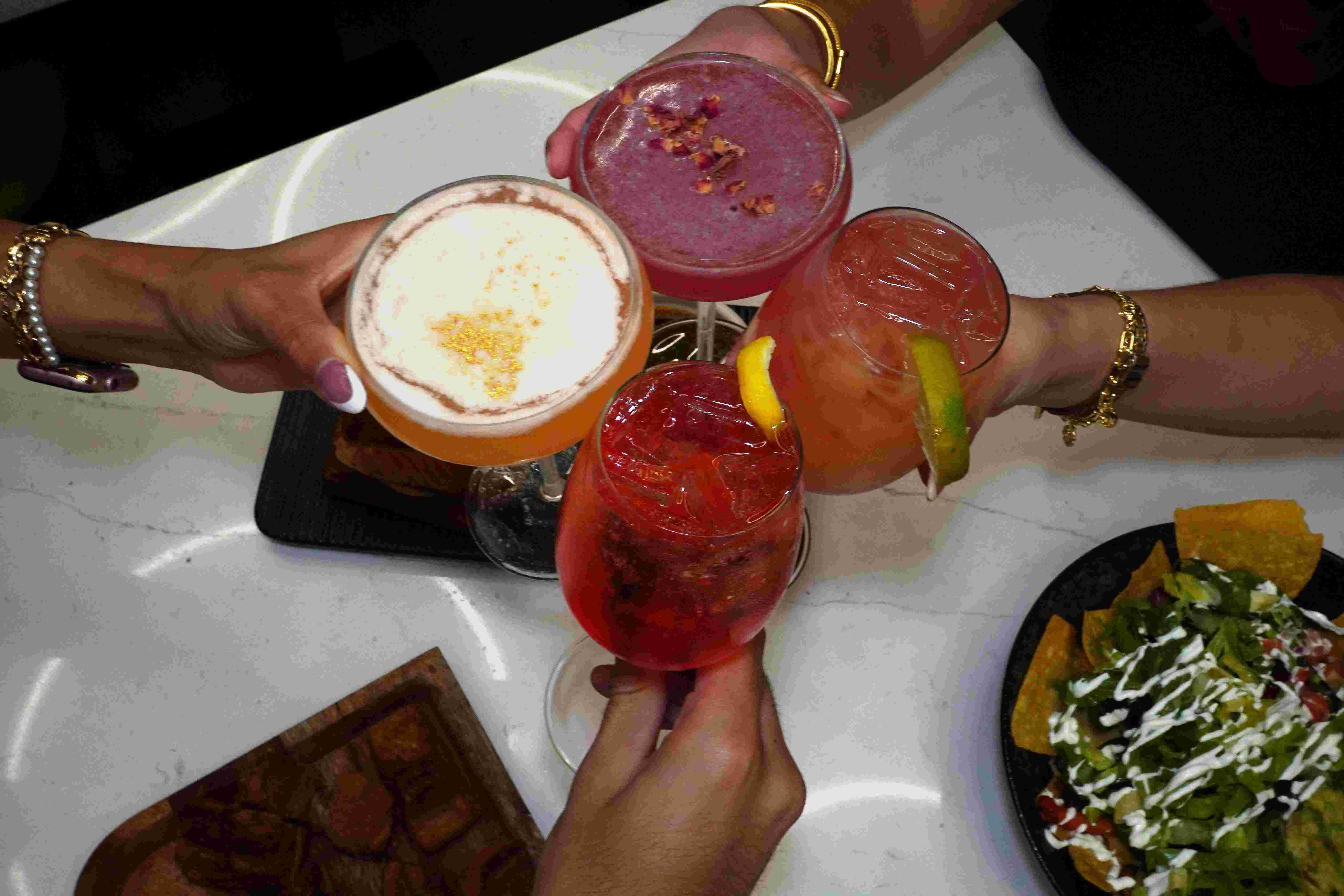Overhead view of hands toasting with various craft cocktails and beverages on a white marble table surface, showing different colored drinks including purple/berry cocktails, creamy drinks with foam, red cocktails, and mixed drinks with lime garnish