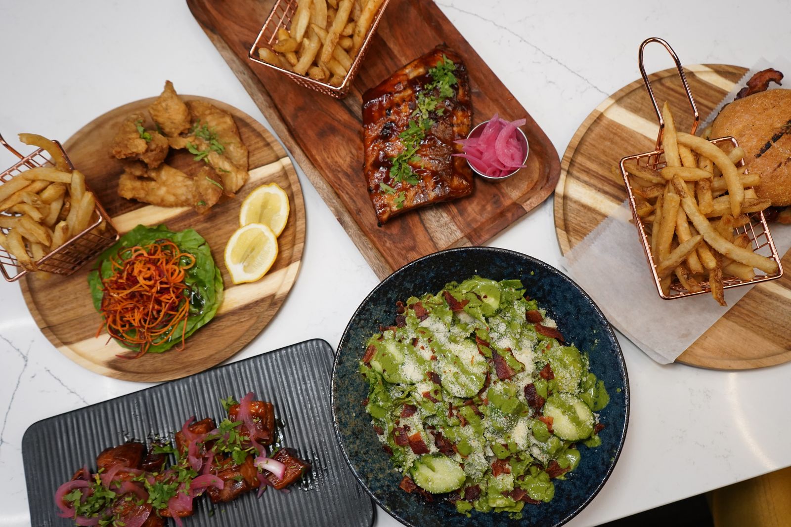 Overhead view of various restaurant dishes on wooden boards and dark plates including fried food with golden fries in copper baskets, glazed ribs with herbs and purple sauce, fresh Brussels sprouts salad with bacon and cheese in blue bowl, and bacon-wrapped appetizers on black plate