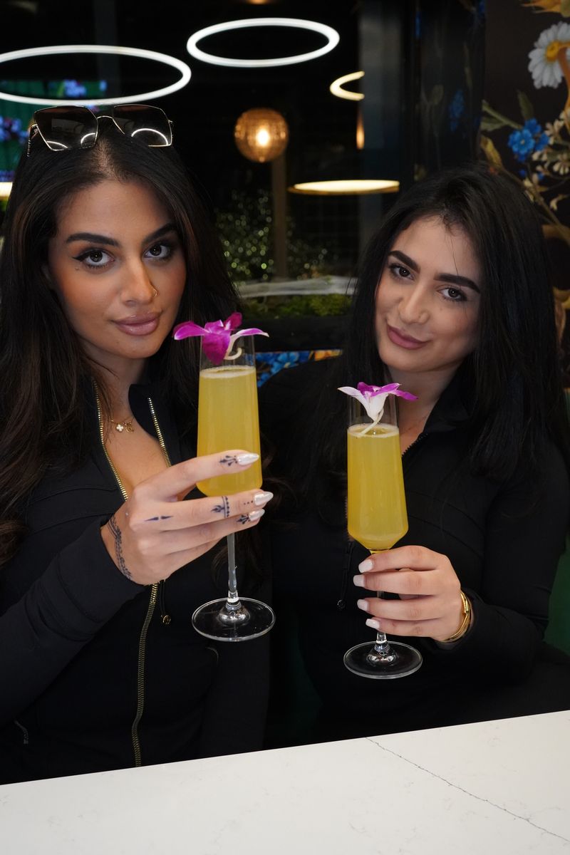 Two women toasting with yellow cocktails featuring purple orchid garnishes in modern restaurant setting with circular pendant lighting and sophisticated dark atmosphere, showcasing craft cocktail experience